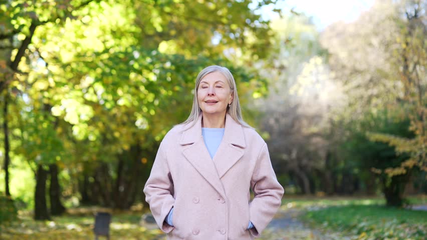 Smiling senior woman in a coat walking through an urban city park, enjoying a pleasant day outdoors surrounded by green trees, feeling relaxed and joyful while appreciating nature and fresh air.