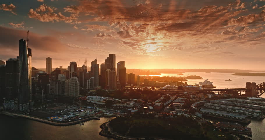 Sydney city skyline at golden hour sunset with Opera House and Harbour Bridge bathed in warm dramatic light, reflecting across the harbor for a vibrant evening cityscape panorama. Aerial drone flight