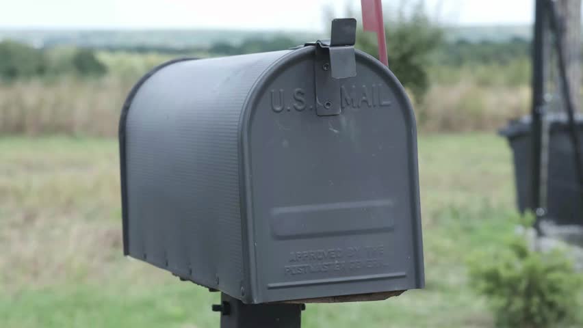 Black U.S. Mail mailbox with a red flag in a rural setting, standing against a blurred background of green fields and trees, symbolizing traditional postal service and communication