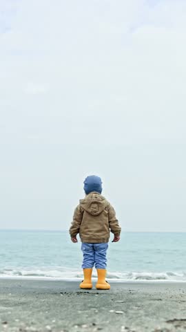 A boy in a jacket and rubber boots walks along the beach near the sea. A little 2 year old boy plays near the sea. The child breathes sea air.