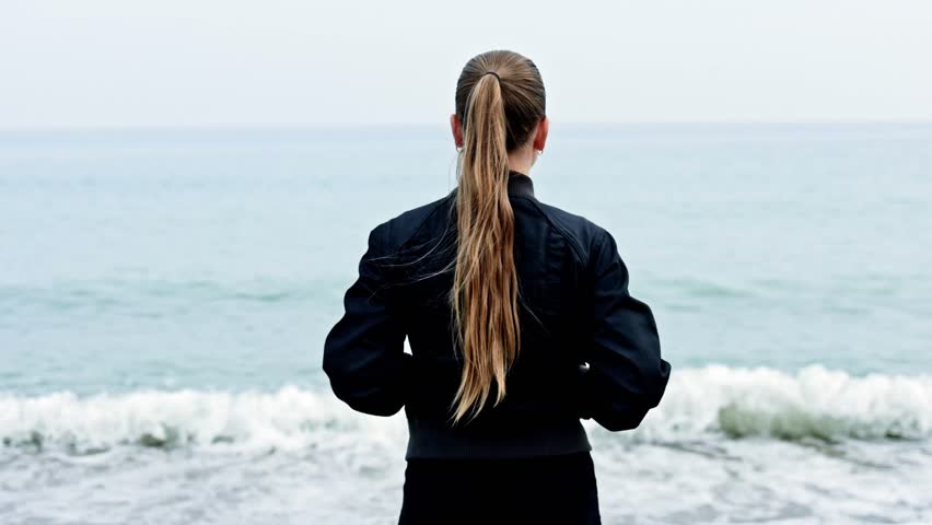 Young woman standing and looking at the sea, view from the back. Woman walking along the seashore. Girl with long hair walking near the ocean in autumn.