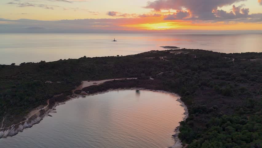 Aerial view of a secluded bay on a wooded peninsula with a distant lighthouse at sunset