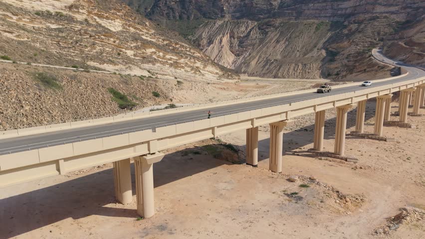 Aerial view of vehicles traveling across a concrete viaduct bridge over a dry valley in the mountainous terrain of Iran.