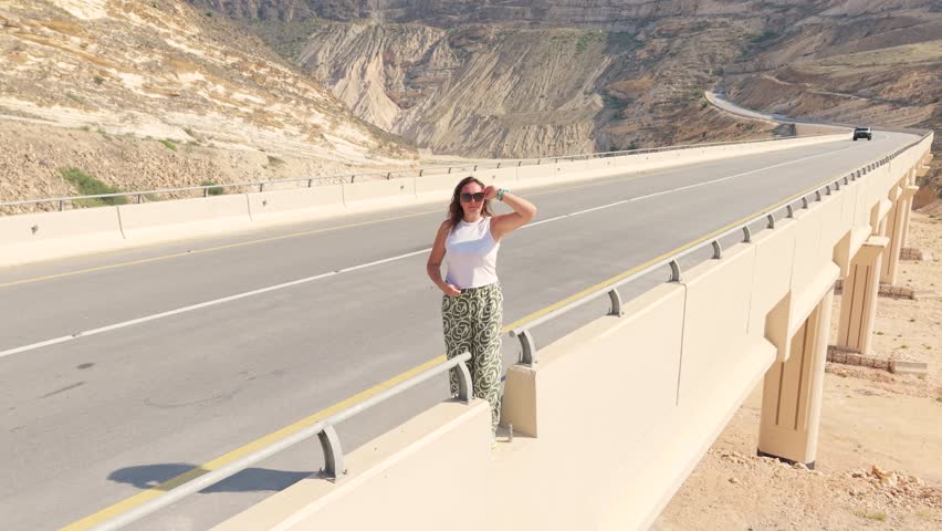 Aerial view of a young woman standing on a concrete bridge spanning a dry valley with vehicles driving on the highway in Iran.