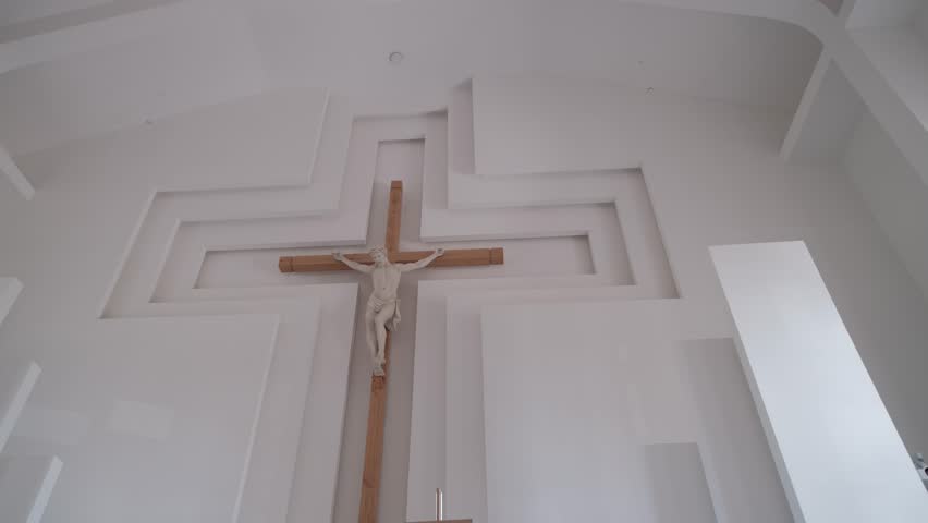 Statue of Jesus Christ on a wooden crucifix mounted against a modern geometric white church wall. Low angle shot.