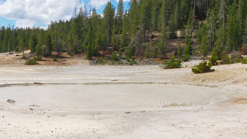 Bubbling geothermal mud pool in hydrothermal basin surrounded by forest hills in Yellowstone National Park, hydrothermal basin landscape.
