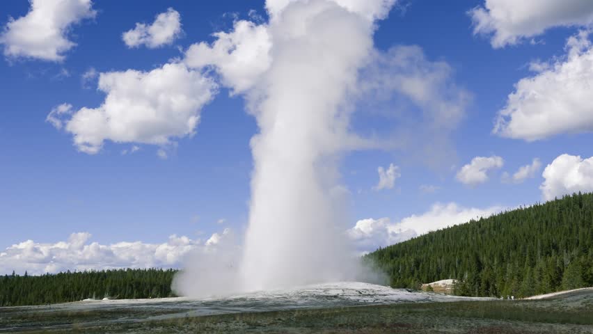 Old Faithful geyser erupting and shooting boiling water high into blue sky in Yellowstone National Park surrounded by forested hills and geothermal landscape
