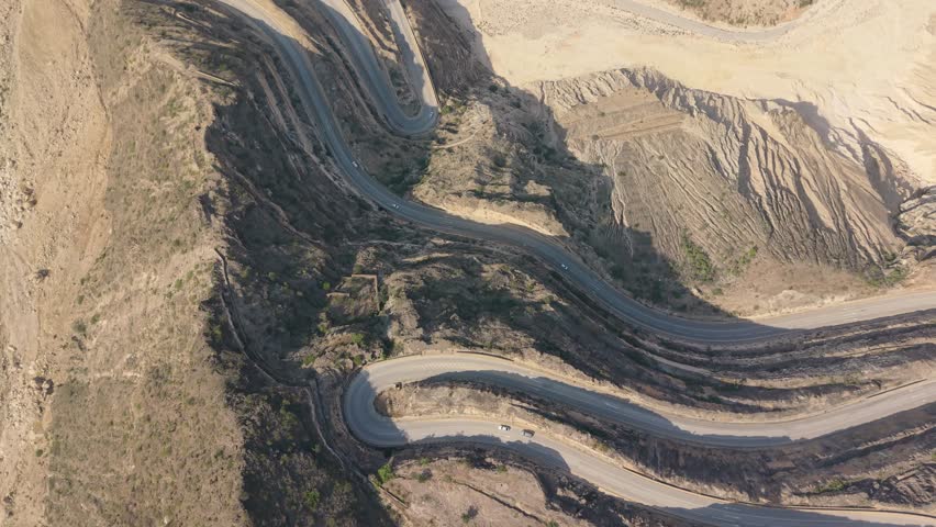 Aerial view of vehicles traveling along a winding mountain road with sharp hairpin turns through an arid landscape in Iran.