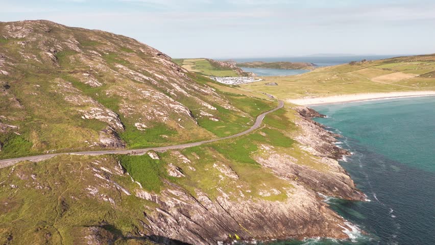 Winding road traces the dramatic coastline of Ireland. Green hills slope to a pristine white sand beach and turquoise Atlantic Ocean. A distant village rests under a bright summer sky.