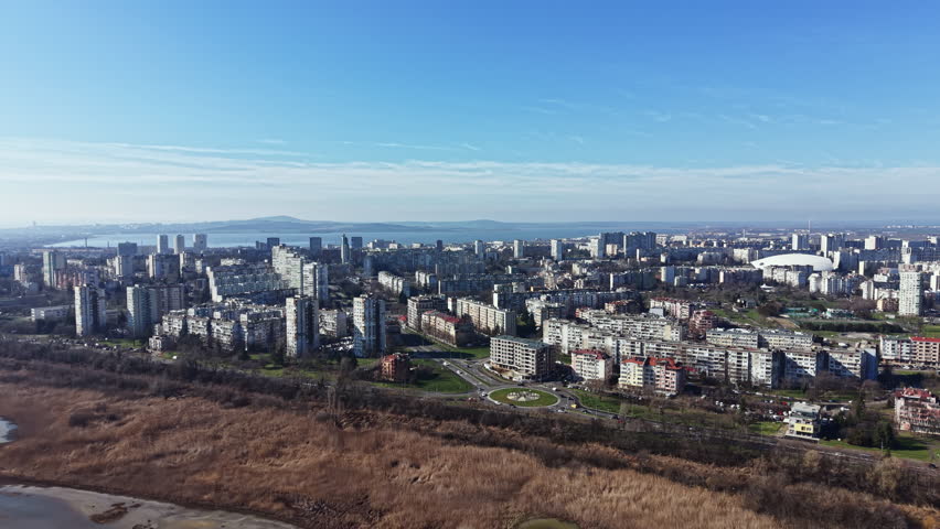 Tall buildings stretch across the city with open land in the foreground. The horizon shows water and a few clouds. It is daytime with sunlight shining.