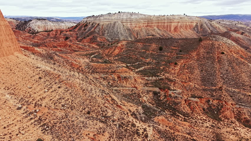 Wide view of a canyon with steep sides and dry ground showing layers of rocks. The landscape is mostly barren with some scattered trees and shrubs.