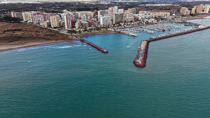 Waterfront buildings line the coast with a harbor filled with boats. Waves crash gently on the shore while a small boat moves toward the harbor entrance in a seaside town.