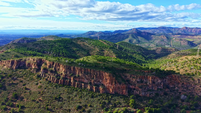 Green hills and rocky cliffs cover a vast landscape under a clear blue sky. The hills rise and fall in the distance, while power lines run along the ridges.