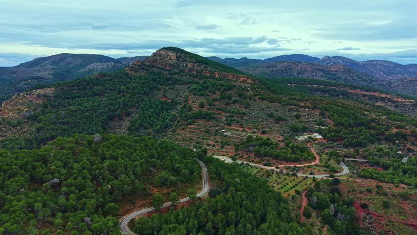 A large green landscape shows rolling hills and trees under a cloudy sky. A winding road cuts through the forest and connects small clearings with distant mountains.