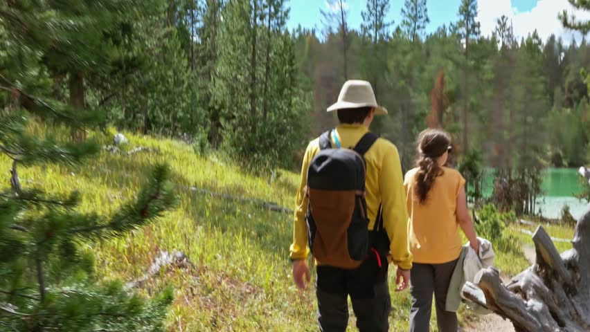 Hikers wearing backpacks walk along a forest trail toward a turquoise lake surrounded by pine trees in Yellowstone National Park, Travel tourism, Outdoor recreation
