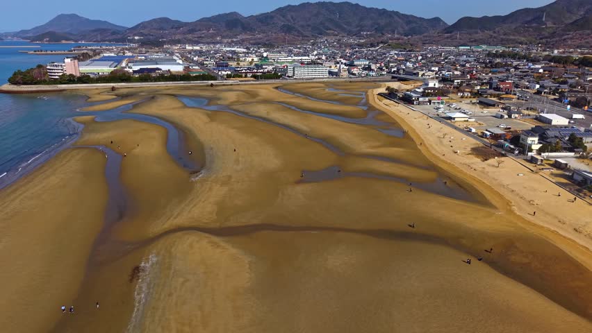 Aerial drone view of Chichibugahama Beach and coastal town in Kagawa, Japan. Scenic tidal flats and sand patterns along the coastline with mountains in the background, captured in 4K.