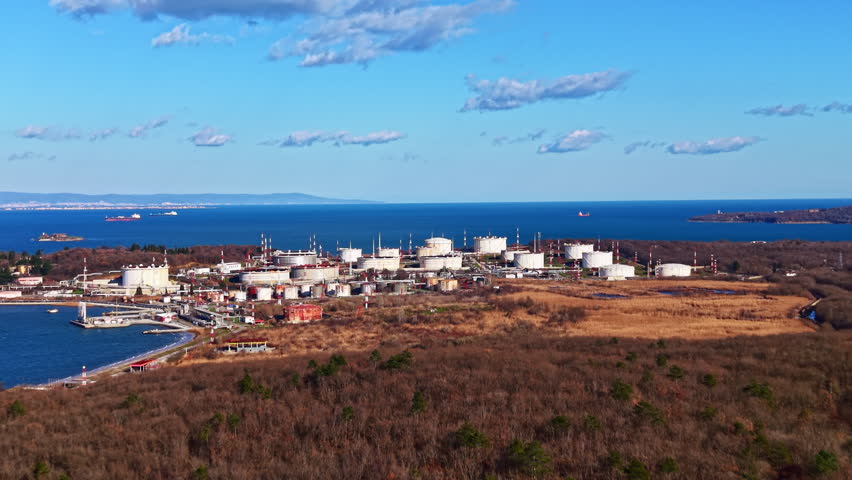 An industrial area features multiple storage tanks located near the shoreline with a view of the ocean and ships in the distance on a clear day.