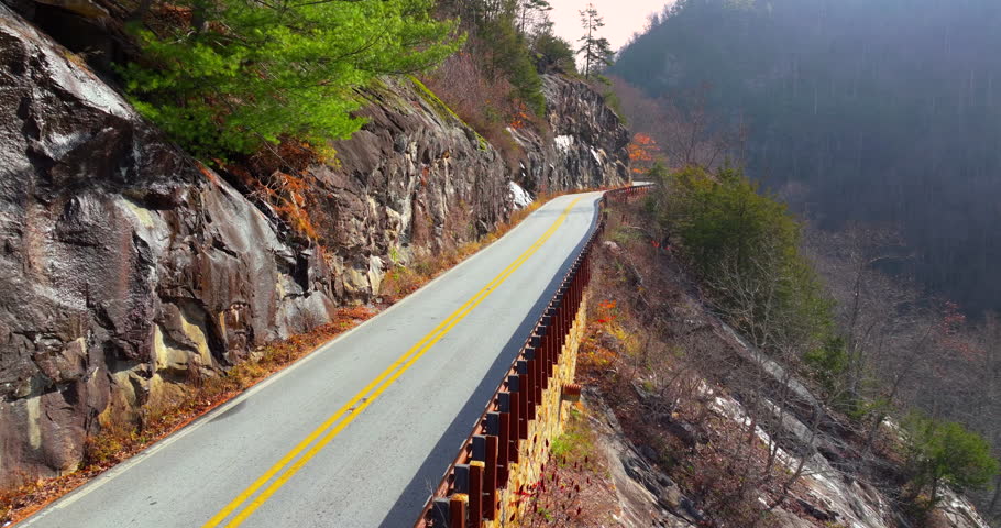 Scenic mountain road in North Carolina Highlands region. Car navigates curves beneath golden trees and steep rocky slopes in fall season.