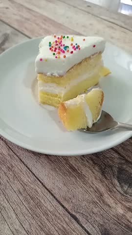 A person eats a slice of vanilla layer cake with white frosting and colorful sprinkles on a white plate over a wooden table.