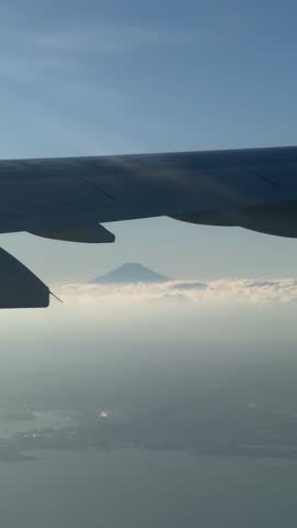 Dramatic aerial silhouette of Mount Fuji at sunset, viewed from an airplane window. The iconic volcanic peak stands out against a vibrant orange and purple sky above a sea of clouds.