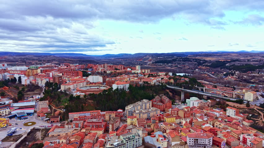 A wide view of a city shows red rooftops and buildings. Mountains rise in the background under a cloudy sky. The scene captures urban life and nature together.