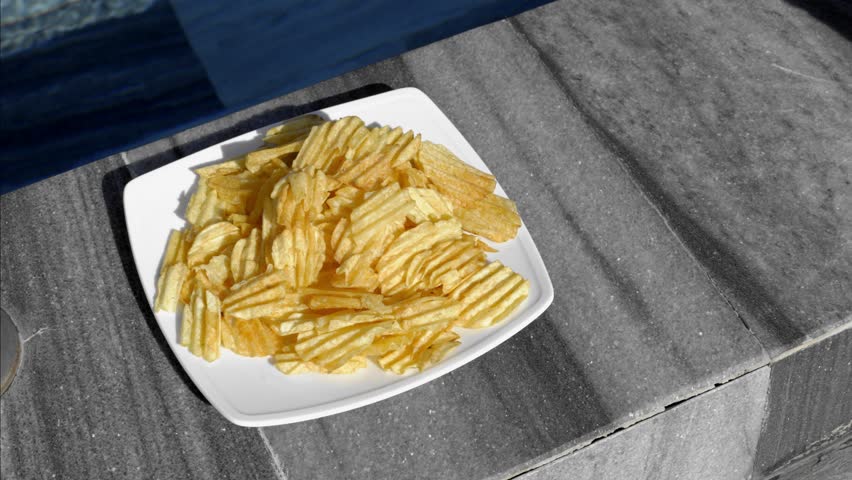 Slow motion static close up of hand picking potato chips from white plate on outdoor stone ledge in sunlight daytime summer setting
