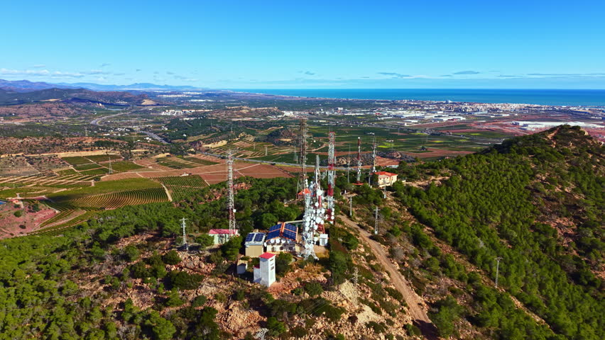 Telecommunication equipment is placed on a high point surrounded by green trees and fields. The sea and city are visible far away under a clear blue sky.