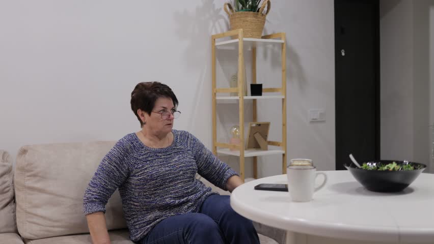 Elderly woman enjoying leisure time at home, alternating between watching television and using her mobile phone