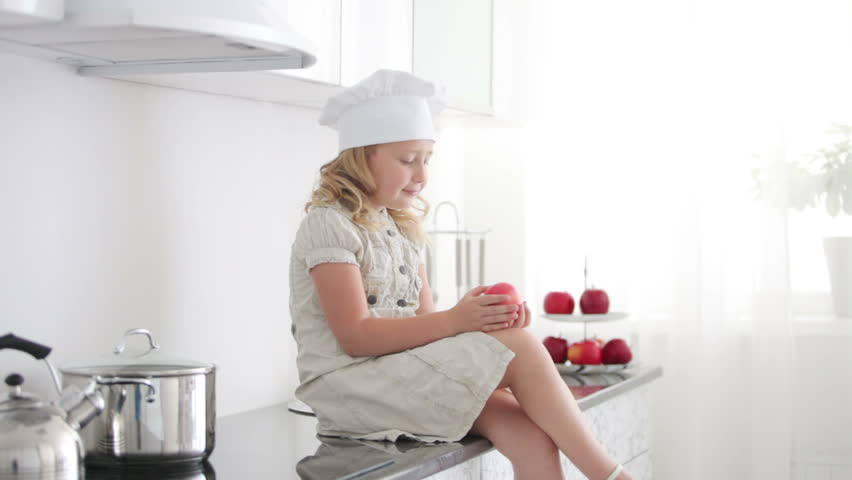 Little girl chef is sitting at the kitchen table and eating an apple
