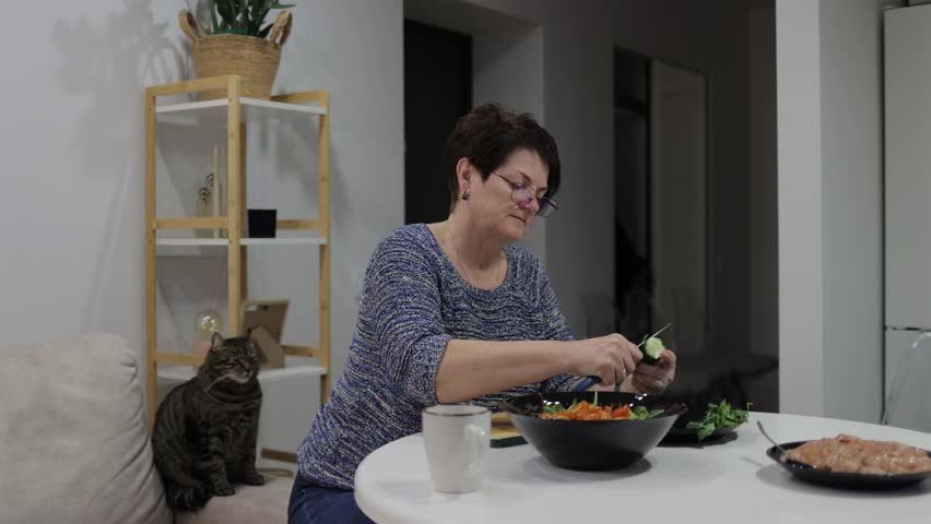 Mature woman carefully slicing fresh cucumber for salad while her tabby cat sits nearby, quietly observing kitchen preparation with peaceful domesticity