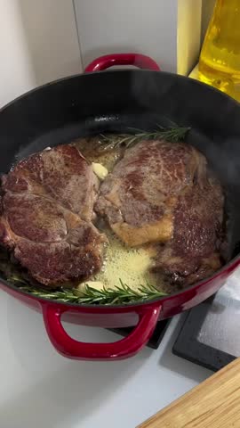 Close-up of beef steak frying in butter with garlic and rosemary in a pan. Chef spooning melted butter over meat while cooking on stove.