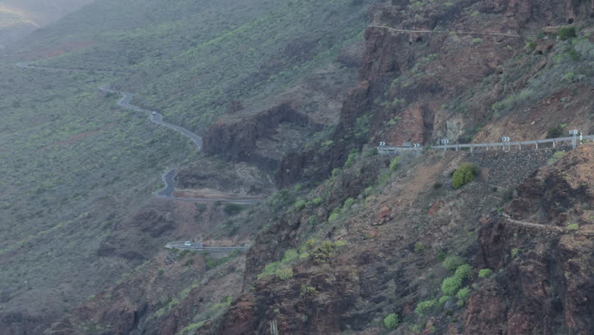 White car drives along a winding mountain cliff road at sunset, passing viewpoints and guardrails in a dry, rocky landscape.