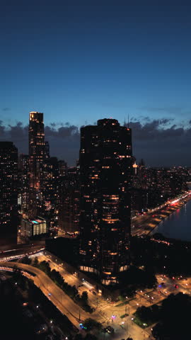 Vertical aerial night view of Chicago Illinois showing downtown skyline, glowing skyscrapers, lakefront roads and flowing traffic lights under deep blue evening sky.