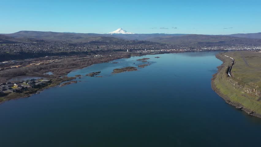 4K aerial drone footage flying above the Columbia River near The Dalles, Oregon, USA with Mount Hood visible in the distance and the town along the shoreline. Scenic Pacific Northwest landscape with river islands and wetlands. A long freight train moves along the riverside railway beneath rocky cliffs, highlighting regional rail transportation and infrastructure. Natural landscape and transportation b roll showing river scenery and rail movement.