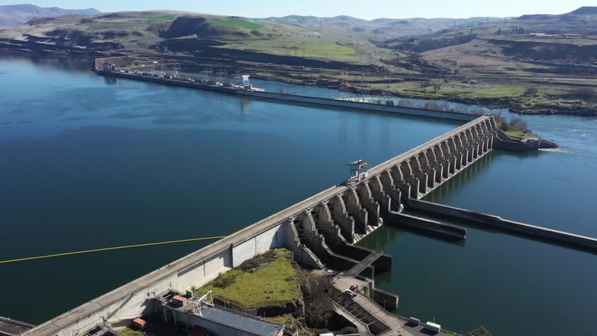 4K aerial drone footage of The Dalles Dam hydroelectric facility on the Columbia River in The Dalles, Oregon, USA. Massive concrete dam structure generating renewable hydroelectric power and controlling water flow along the river. Large scale energy infrastructure supplying electricity to the regional power grid across the Pacific Northwest. Scenic aerial view of the Columbia River and surrounding landscape highlighting renewable energy production and modern utility engineering, great for b-roll