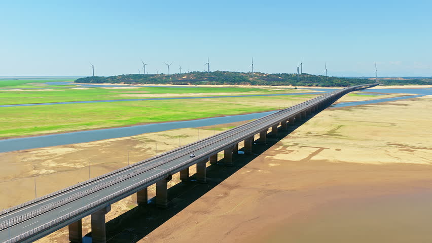 Aerial view of long highway bridge crossing vast wetland and dry riverbed in China, with wind energy turbine on the horizon.
