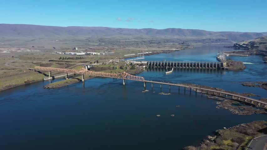 4K aerial drone footage showing The Dalles Dam hydroelectric facility and a large steel truss bridge crossing the Columbia River in The Dalles, Oregon, USA. Major infrastructure combining renewable hydroelectric power generation and regional transportation crossing the river. Scenic aerial view of dam spillways, river water flow, and bridge structure connecting both sides of the Columbia River within the Pacific Northwest landscape.
