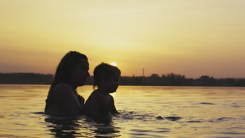 Mom with the baby she holds in her arms playing with her daughter and splashing in the lake against background of sunset
