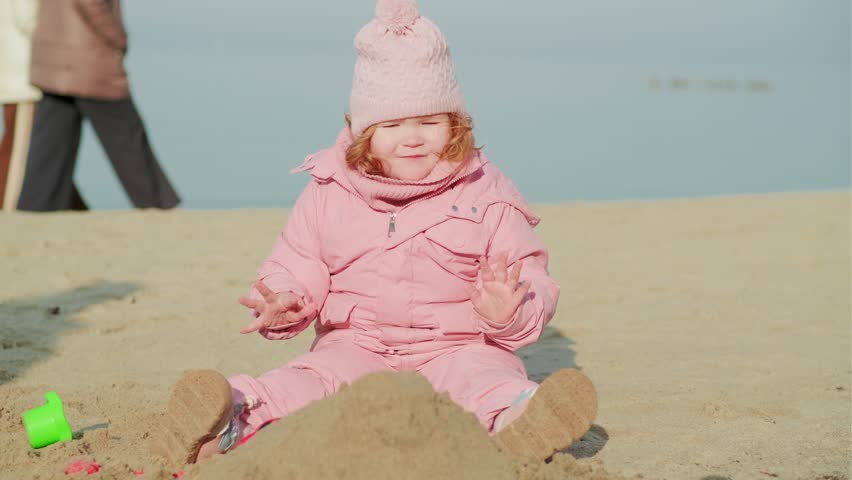 Child girl wearing pink snowsuit sitting on beach sand by sea. Cute toddler playing outdoors in cold weather. Winter seaside adventure for kid. Childhood leisure and family travel concept.