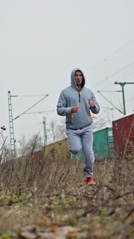 White Man Training Trail Run Near Railway Tracks, Hoodie And Bright Running Shoes, Steady Stride Over Rough Ground, Outdoor Endurance Session With Focused Pace And Gritty Urban Nature Backdrop
