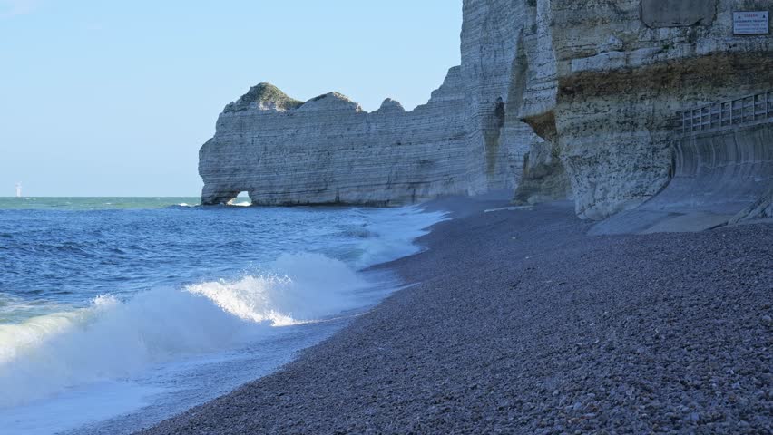 Waves reaching pebble beach below famous Etretat cliffs along Normandy coast. Natural coastal landscape with white chalk formations and Atlantic sea creating iconic France scenery