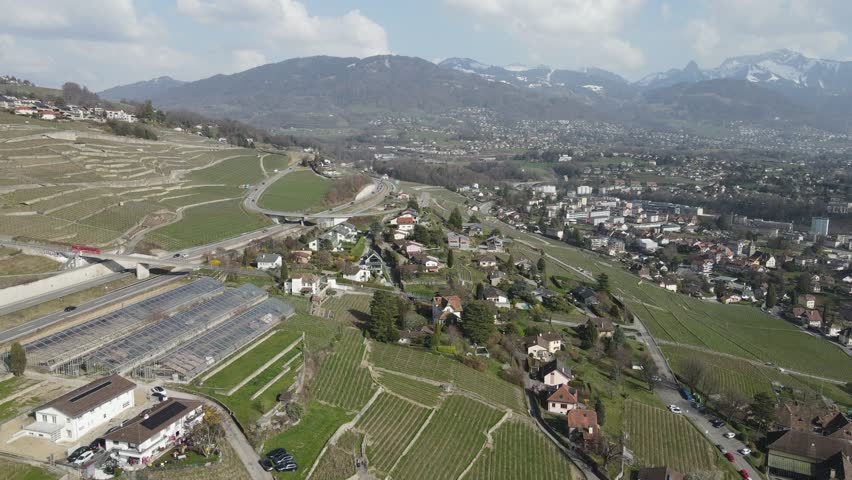 Aerial drone view above the Lavaux vineyard terraces near Vevey and Corseaux on the shores of Lake Geneva, Switzerland. Scenic alpine landscape with vineyards, residential houses and road traffic, while a hillside funicular descends through the vineyards with the Swiss Alps in the background.
