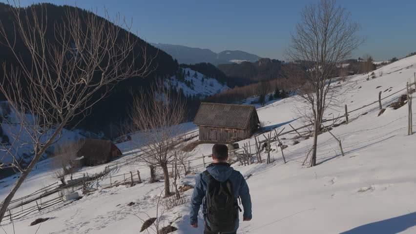 Cinematic tracking shot of a happy hiker walking down a snowy mountain trail among rustic barns at sunset, graded
