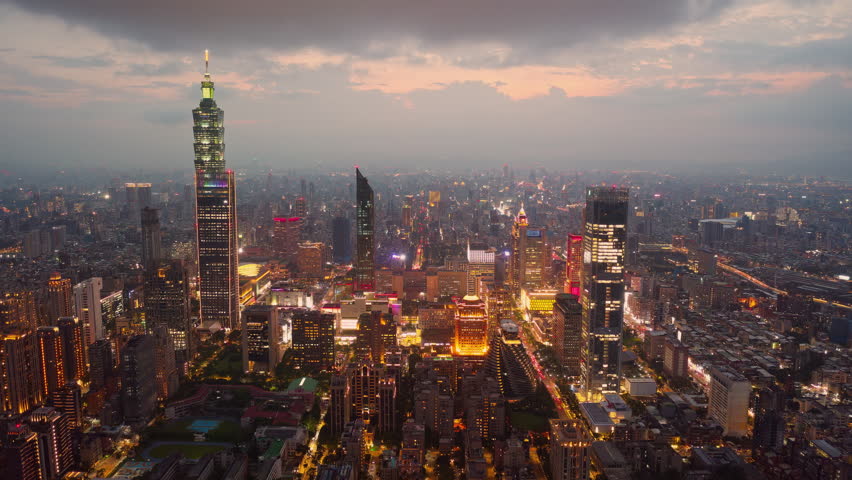 Aerial hyperlapse footage from dusk till night above Downtown Taipei, the vibrant capital city of Taiwan, with landmark 101 Tower standing out amid modern skyscrapers in XinYi Commercial District and city lights dazzling under twilight sky.