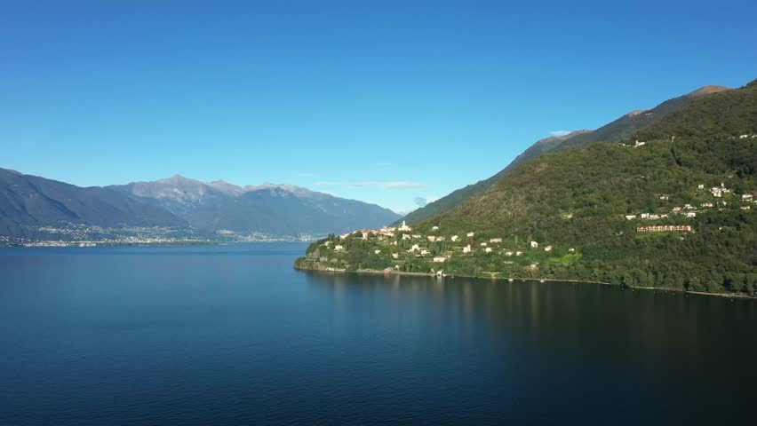 A tranquil view of Lago Maggiore with forested hills and distant mountains under a clear blue sky. Picturesque lakeside villages dot the green slopes.