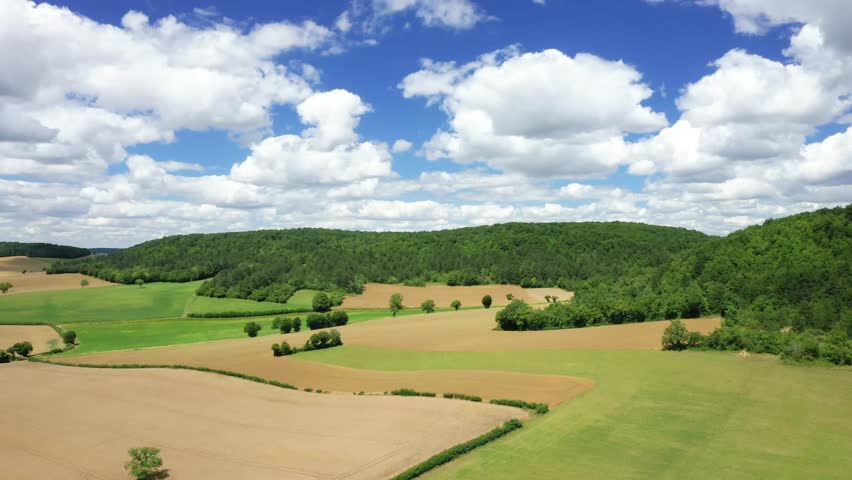Aerial view of rolling fields and lush green forest under a blue sky with scattered clouds in the French countryside during summer.