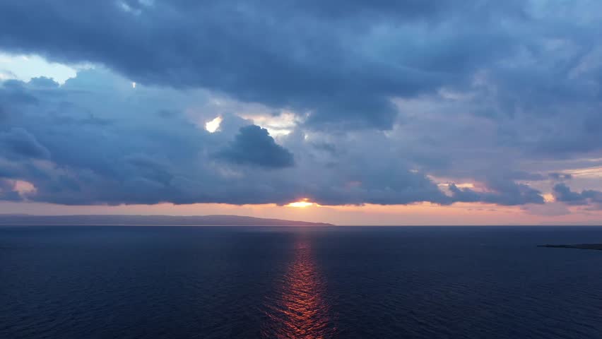 Moody clouds and vibrant sunset colors reflect on the calm sea in Laconia, Greece, creating a serene coastal landscape.