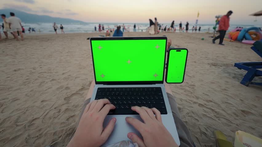 
POV working on laptop with smartphone green screen on beach