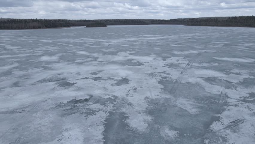 Drone flying over a frozen ice covered lake with a forest in the distance and a cloudy sky