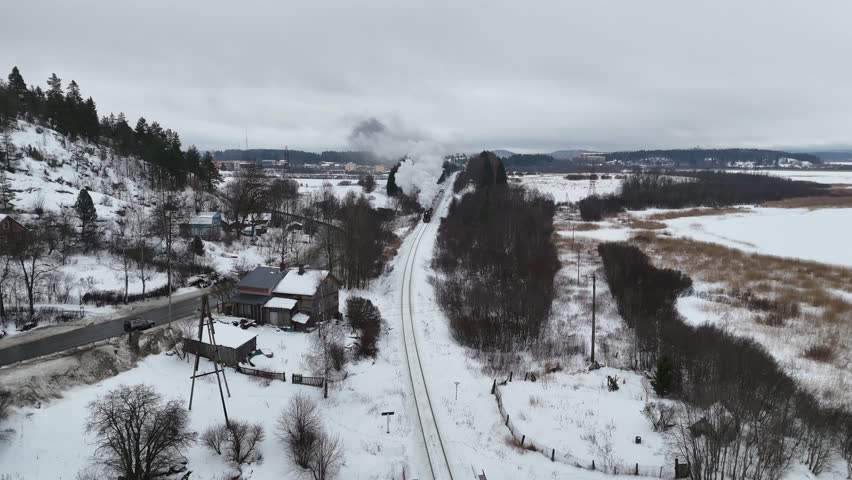 A steam train moves along tracks surrounded by snow and mountains. The train passes houses and fields under a cloudy sky during winter. Smoke rises from the engine as it travels